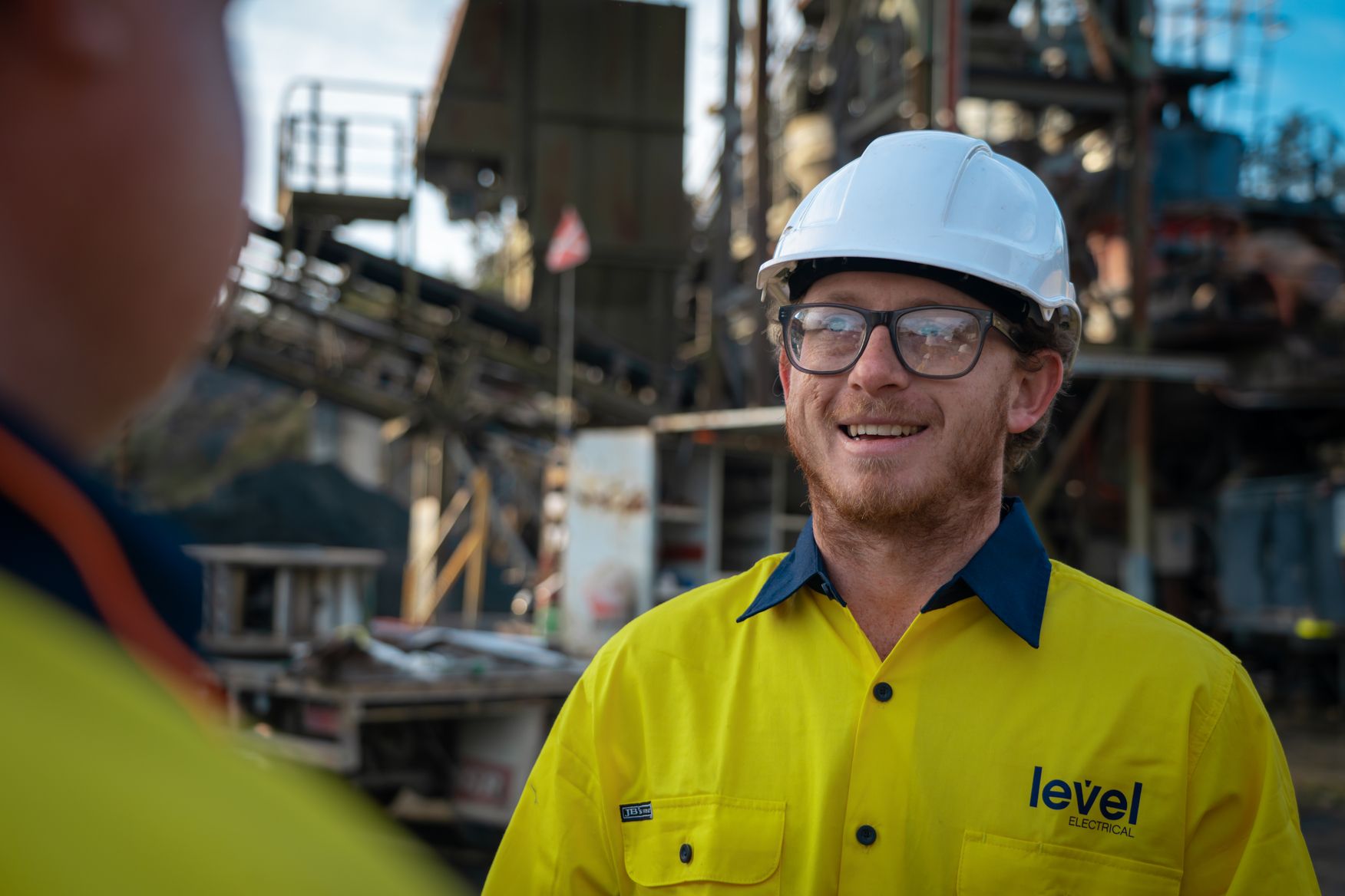 Level Electrical technician wearing safety gear speaking with a colleague on an industrial job site in Australia