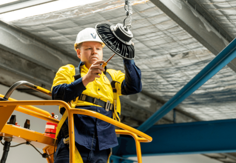 Level electrician installing overhead lighting from a scissor lift on an industrial site in Australia