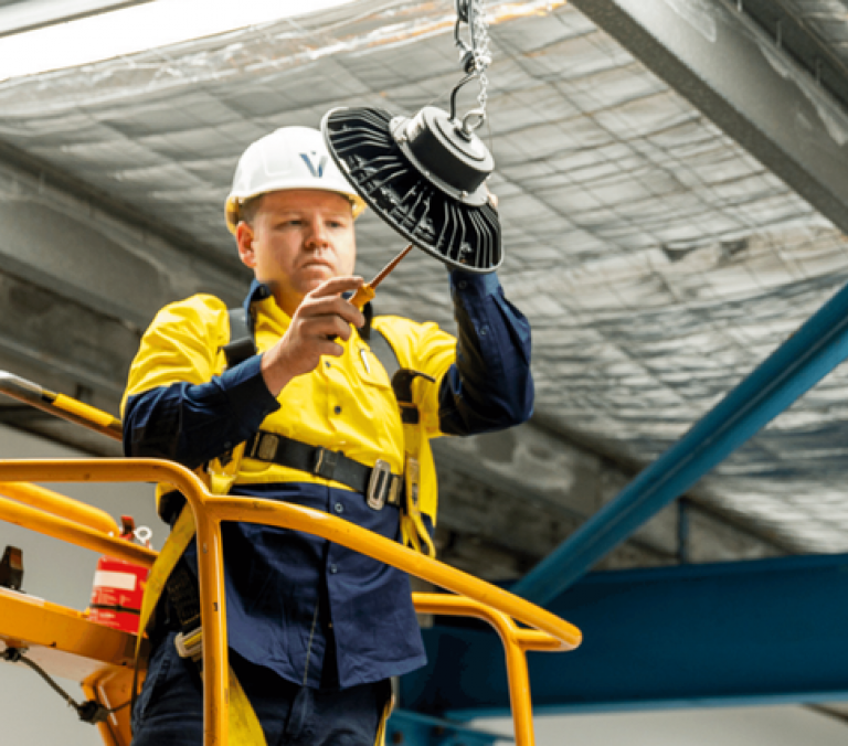 Level electrician installing overhead lighting from a scissor lift on an industrial site in Australia