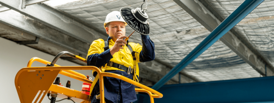 Level electrician installing overhead lighting from a scissor lift on an industrial site in Australia
