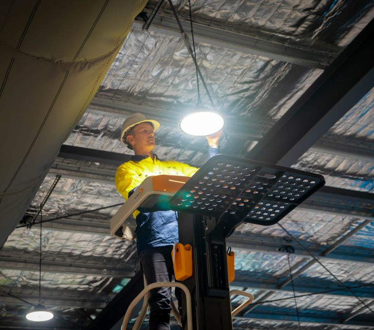 Level electrician installing overhead lighting from an elevated work platform on an industrial site in Australia