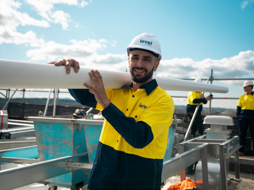 Level plumbing technician carrying pipe during rooftop infrastructure installation on a commercial building in Australia